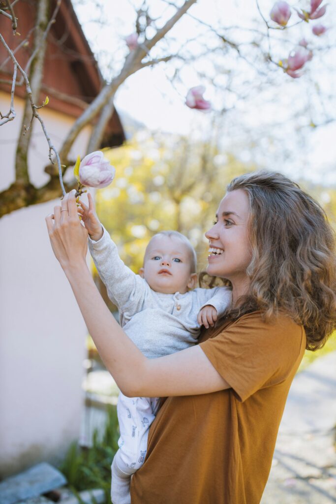A mother happily holds her baby up to reach blooming magnolias in a sunny garden.